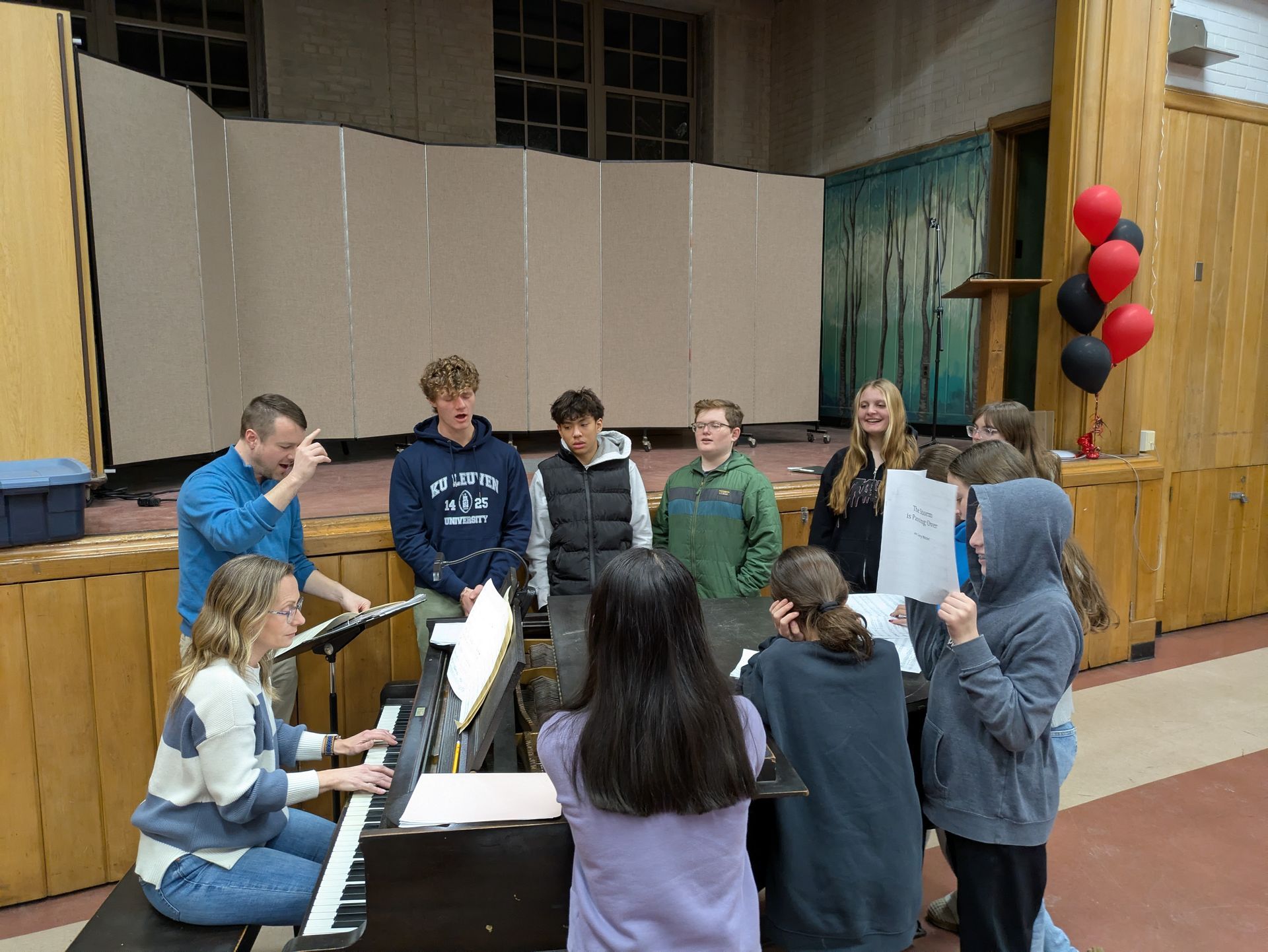 Students gather around a piano during a rehearsal on a stage, with red and black balloons at the side.