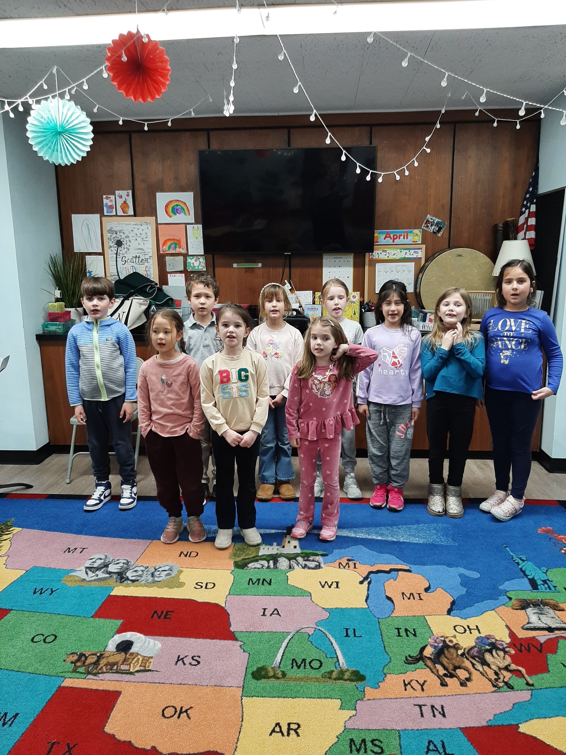 Children standing in a classroom in front of a TV, on a colorful alphabet rug with paper decorations overhead.