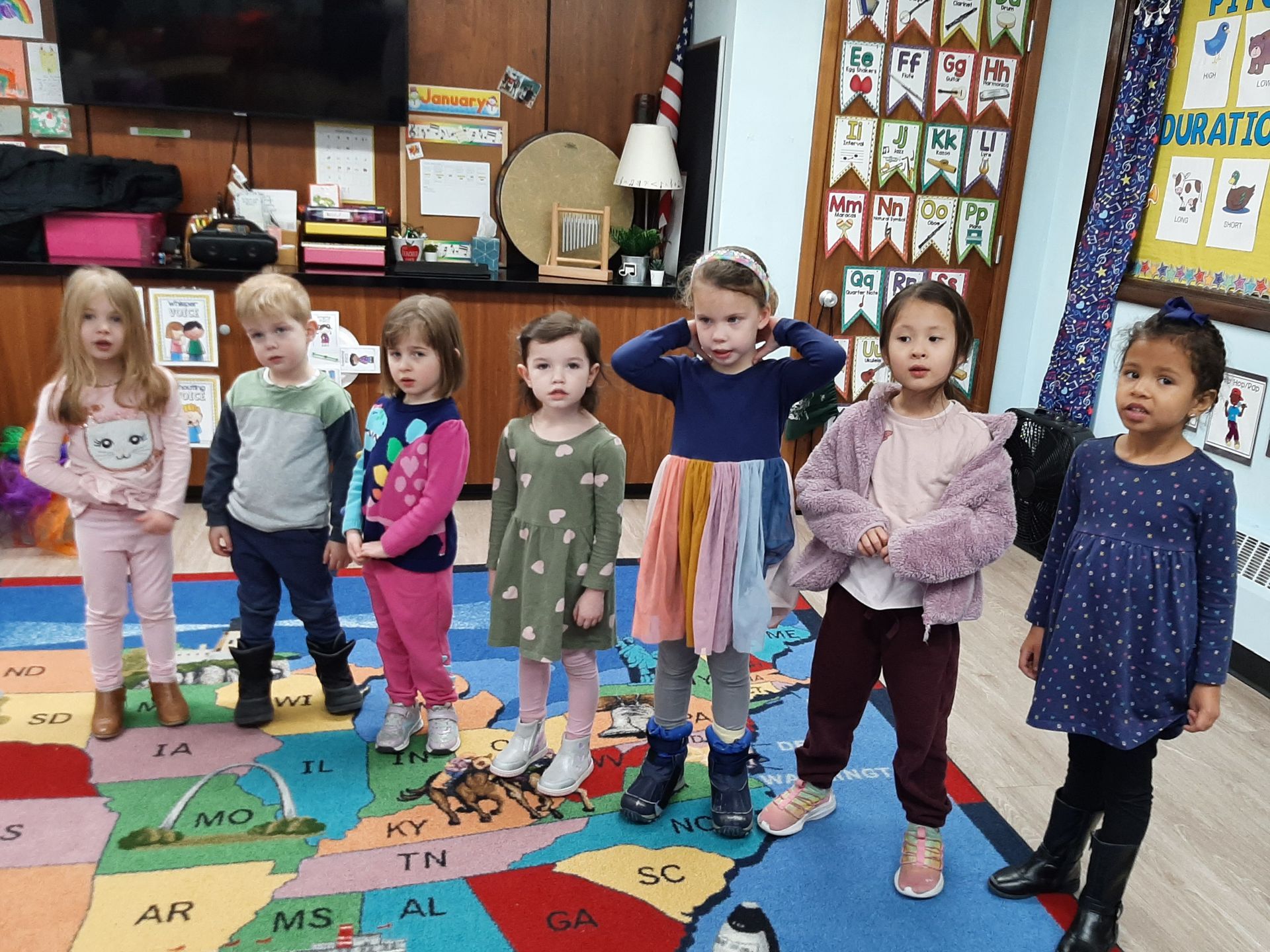 Six children standing on a classroom rug, posing in front of a wall with colorful decorations.