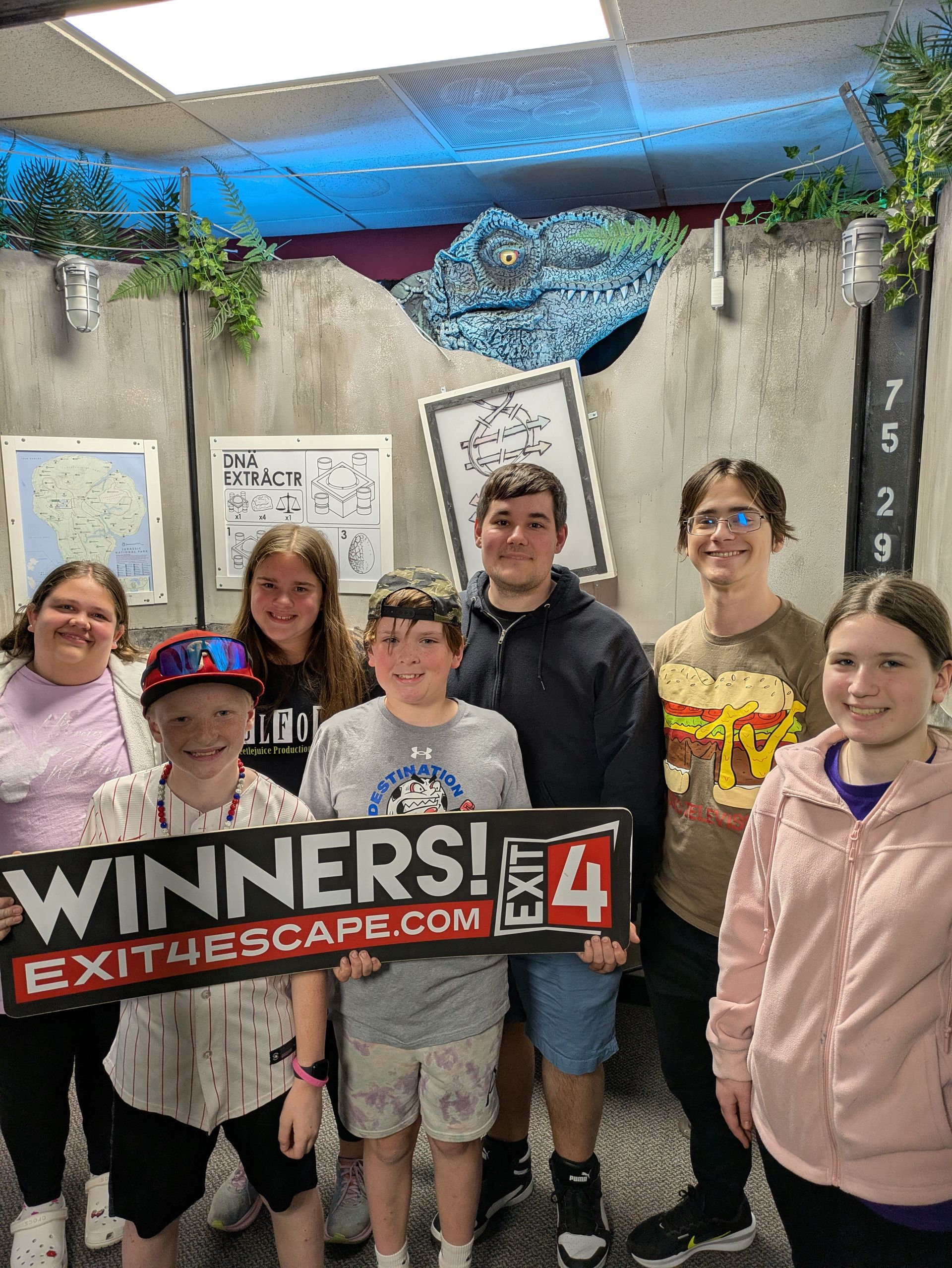Group holding a “Winners” sign in front of an escape room wall, smiling at the camera.