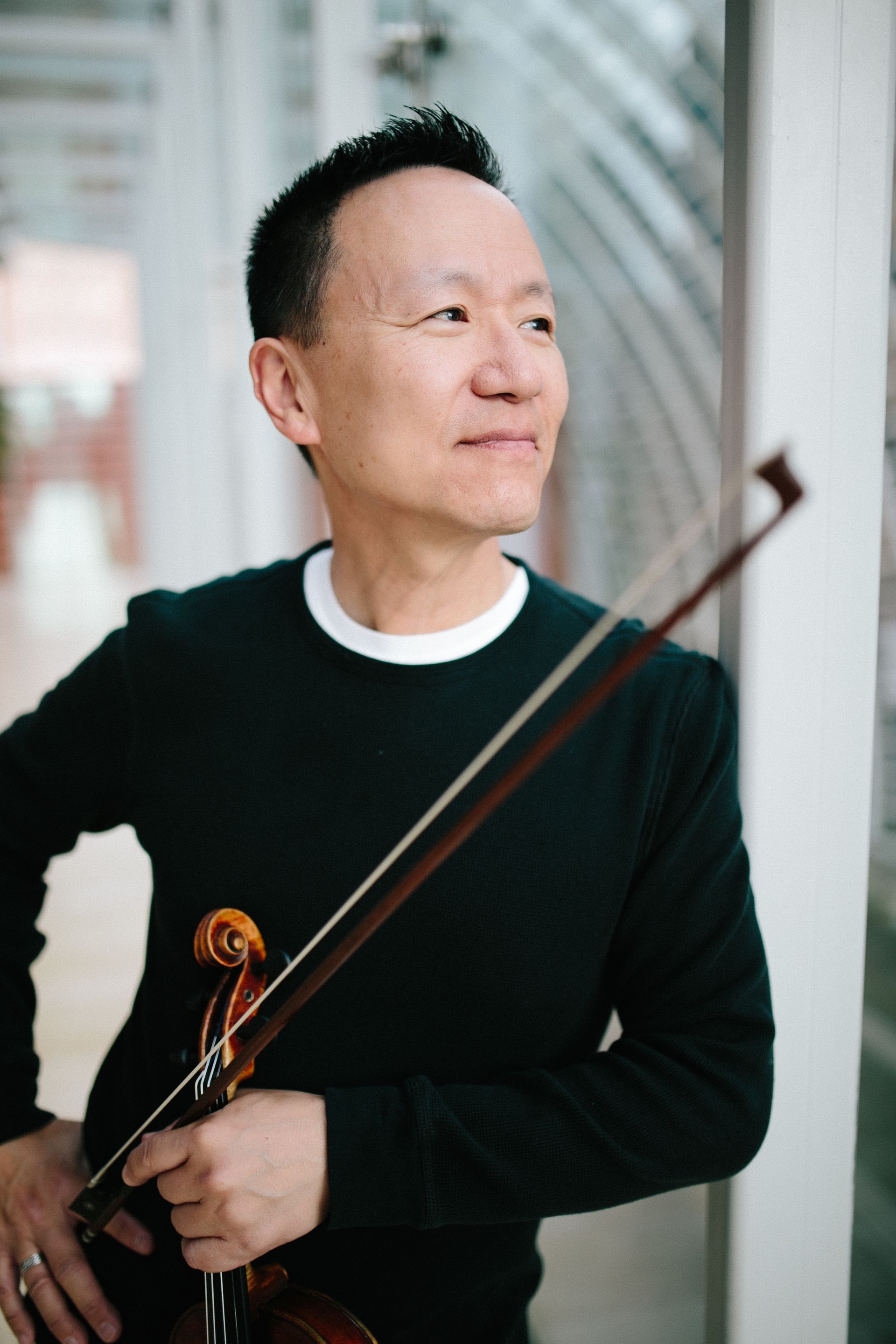 Man holding a violin and bow, wearing a black sweater, looking to the side indoors.