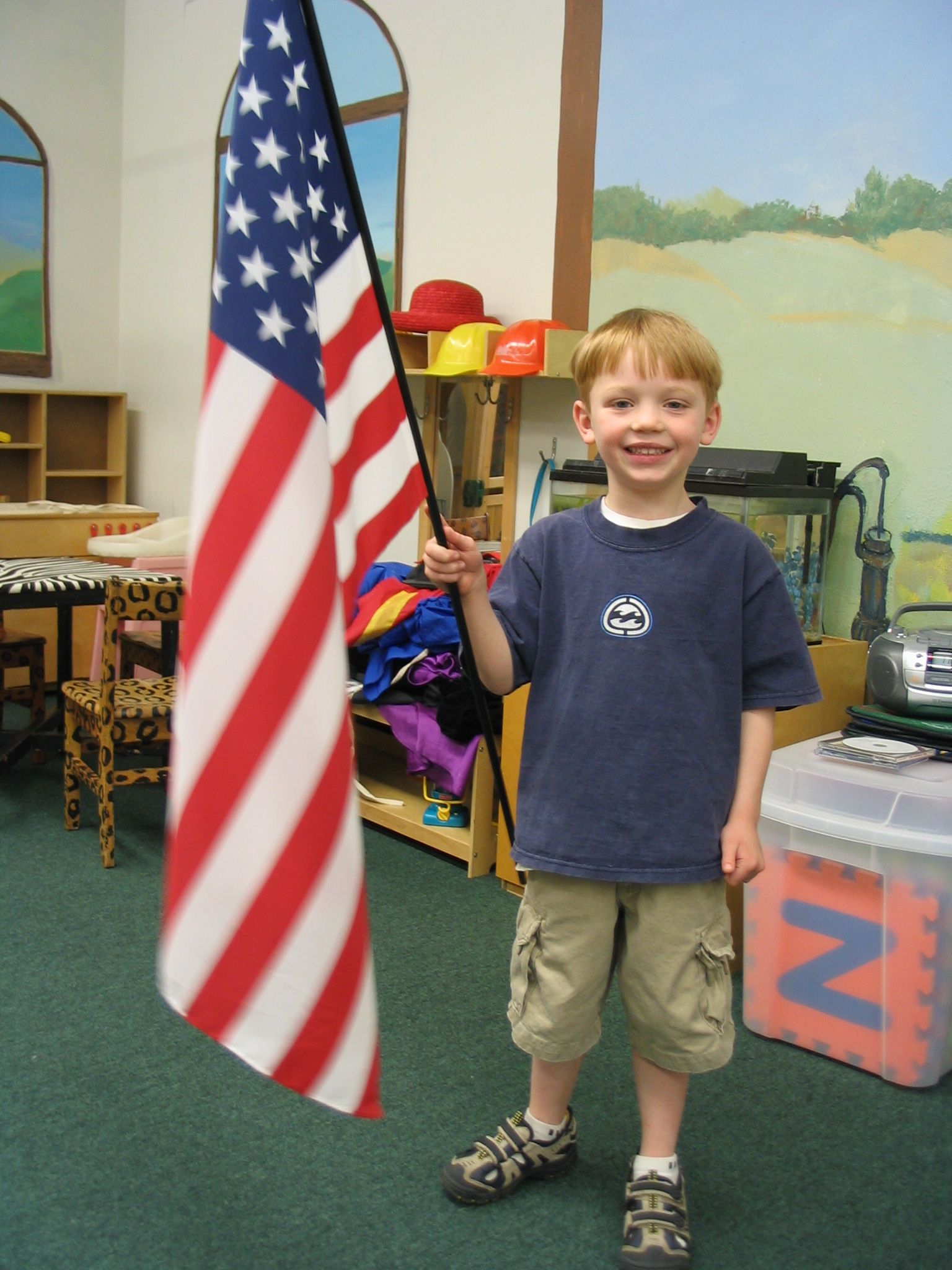 Boy standing indoors beside a large American flag, smiling and holding the flagpole.