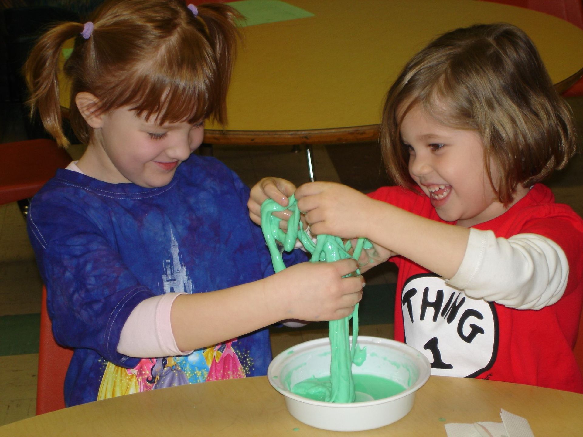 Two children playing with green slime at a table, smiling in a colorful classroom.