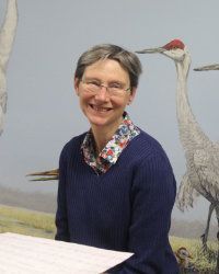 Smiling person in glasses and navy sweater, standing before a mural with cranes.