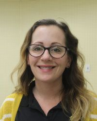 Smiling person with glasses and wavy hair, wearing a yellow cardigan and black shirt against a light wall.