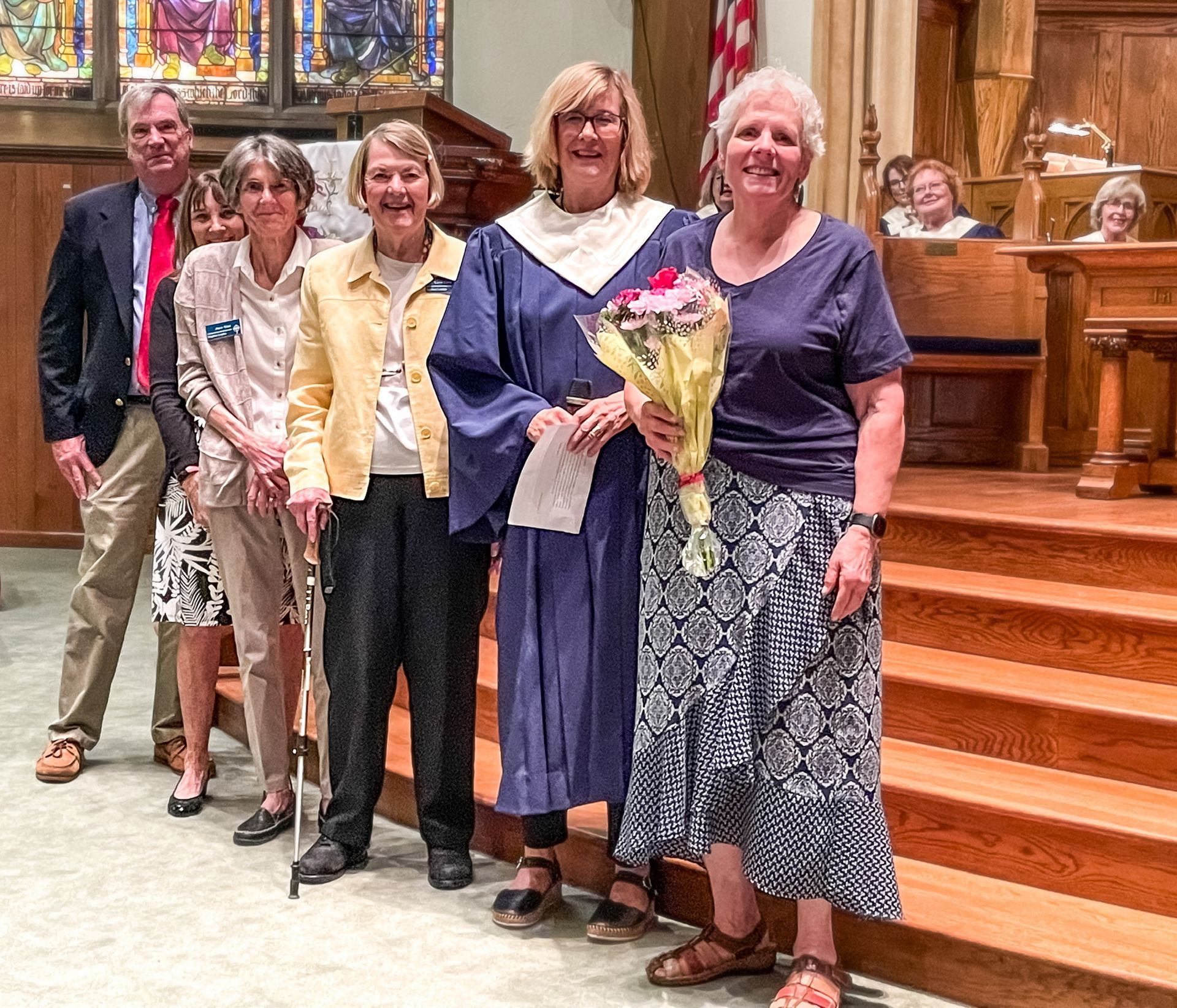 Group of five people posing in a church sanctuary, one holding flowers, with stained glass and wooden pews behind them