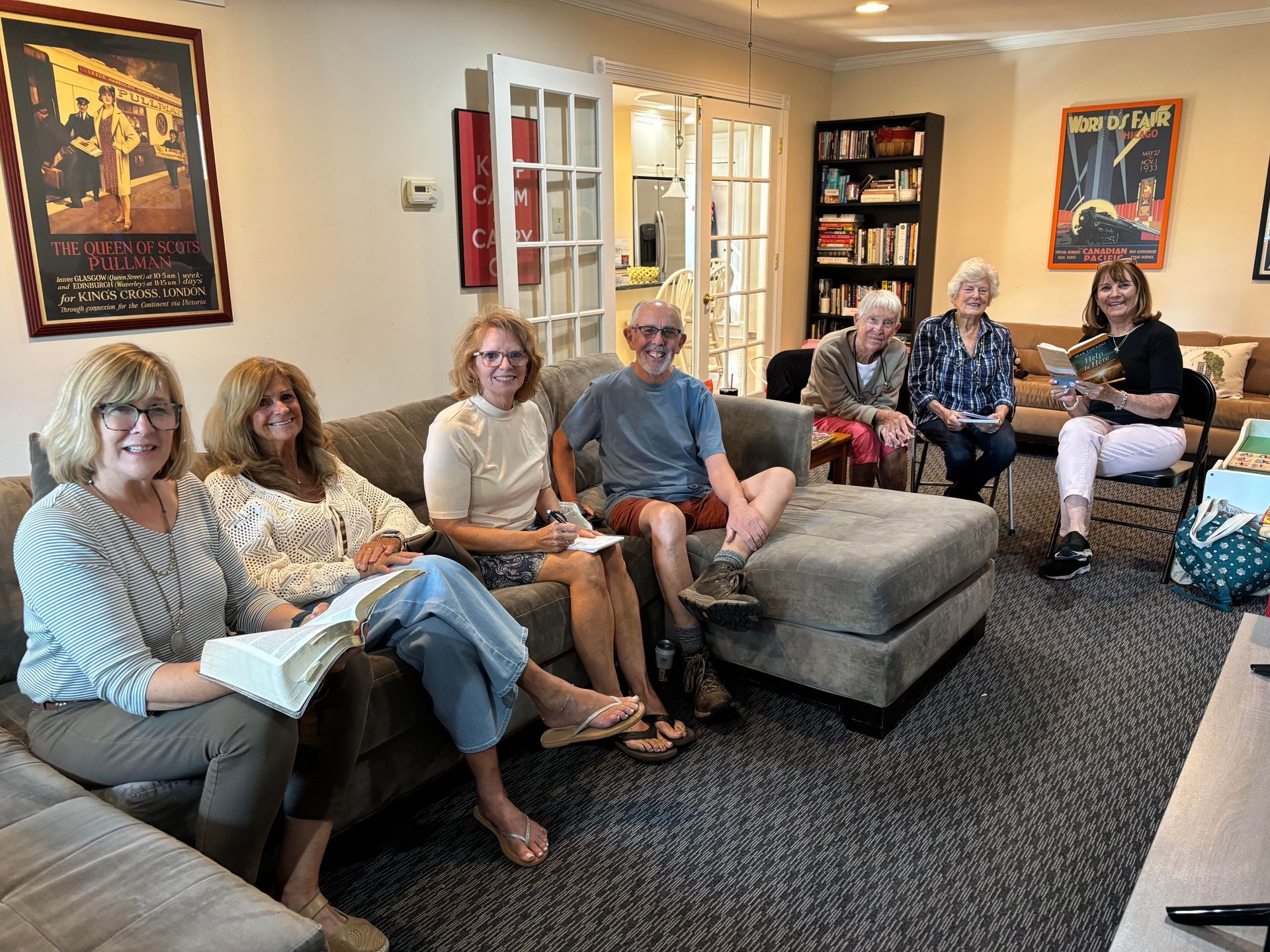 Group of people sitting and chatting in a cozy living room with bookshelves and framed wall art.