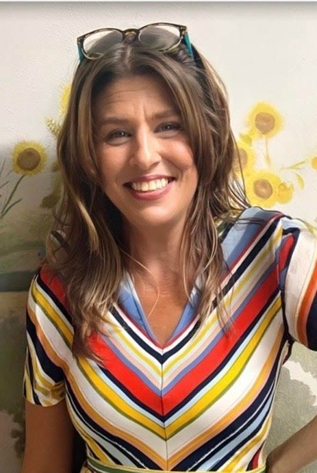 Smiling woman in a striped V-neck top with sunglasses on her head, standing in front of sunflower wall art