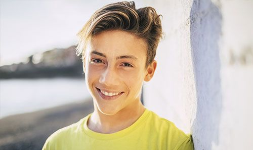 Smiling person in a yellow shirt leaning against a white wall outdoors