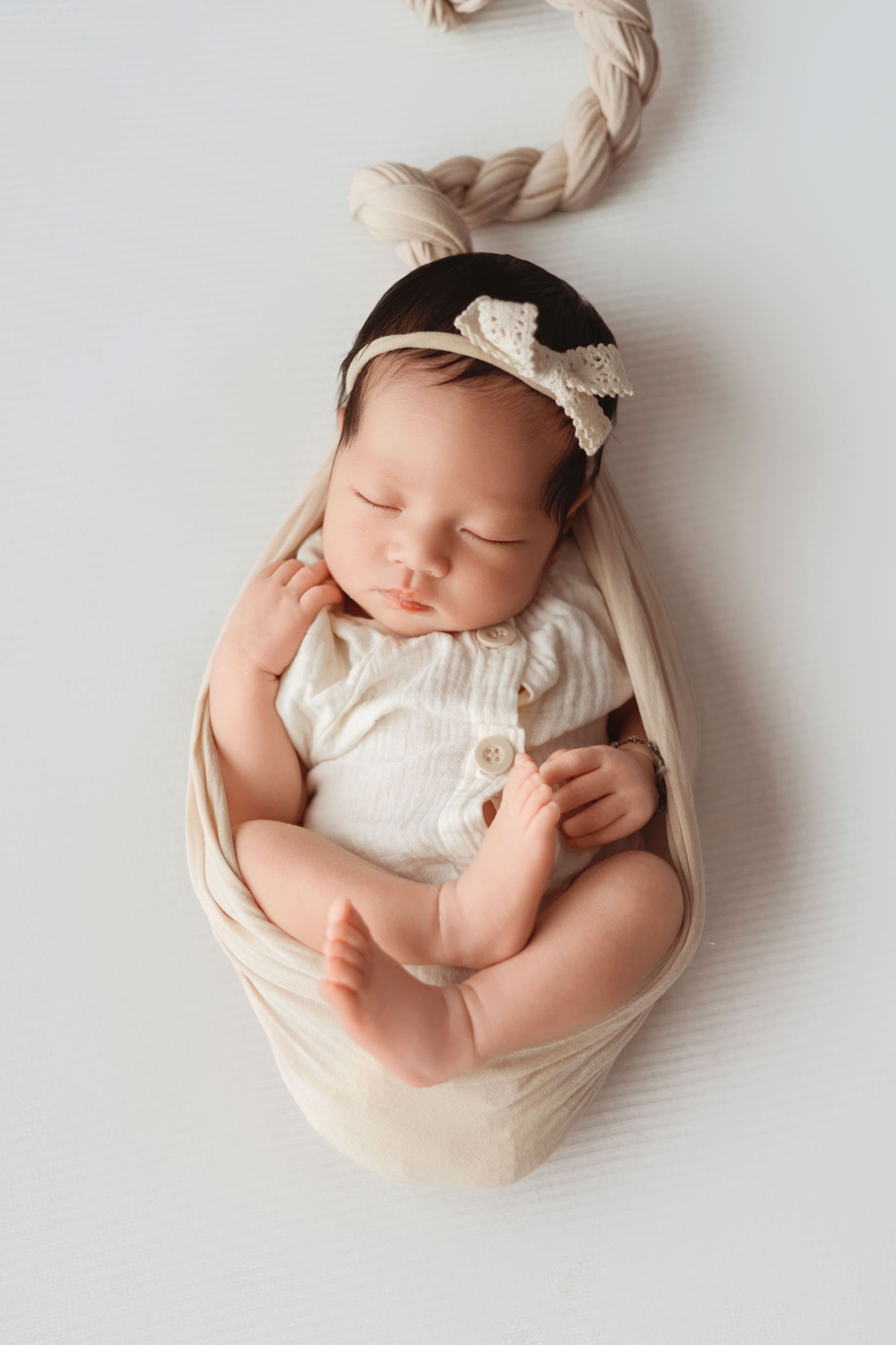 Sleeping baby wrapped in cream blanket with a bow headband, nestled in a hanging basket.