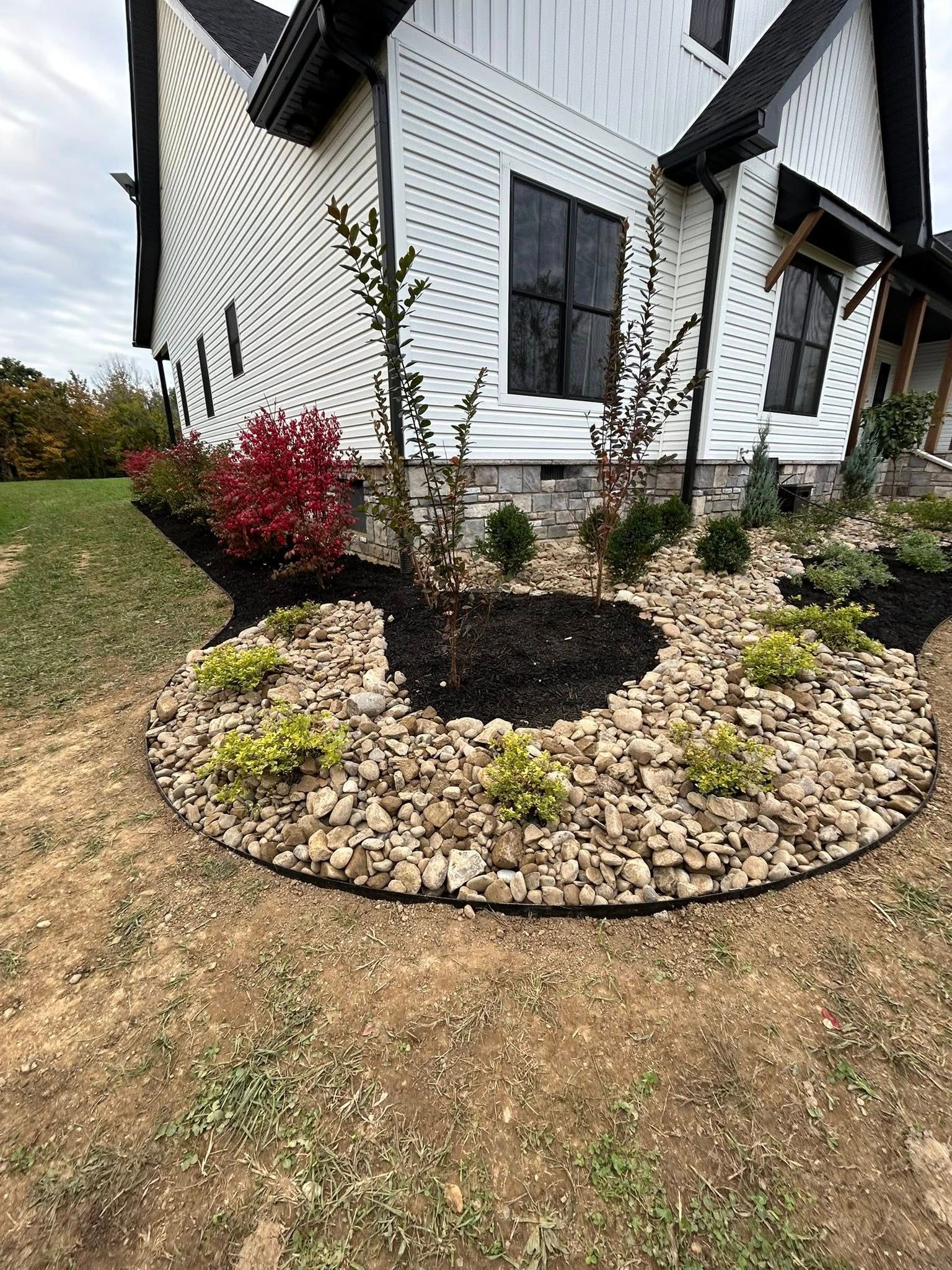 A landscaped garden bed with rocks, mulch, and plants in front of a white house.