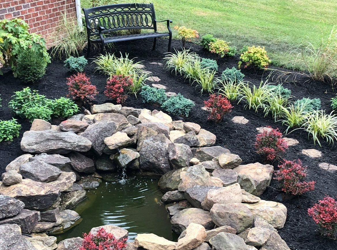 Stone-lined pond in a garden bed with colorful plants, stepping stones, bench, and fresh black mulch.