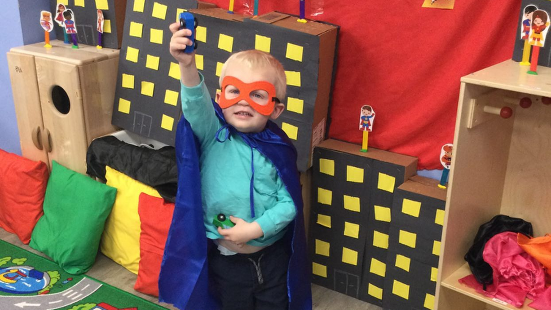 Child in superhero costume, holding toy, in a play setting with city backdrop.