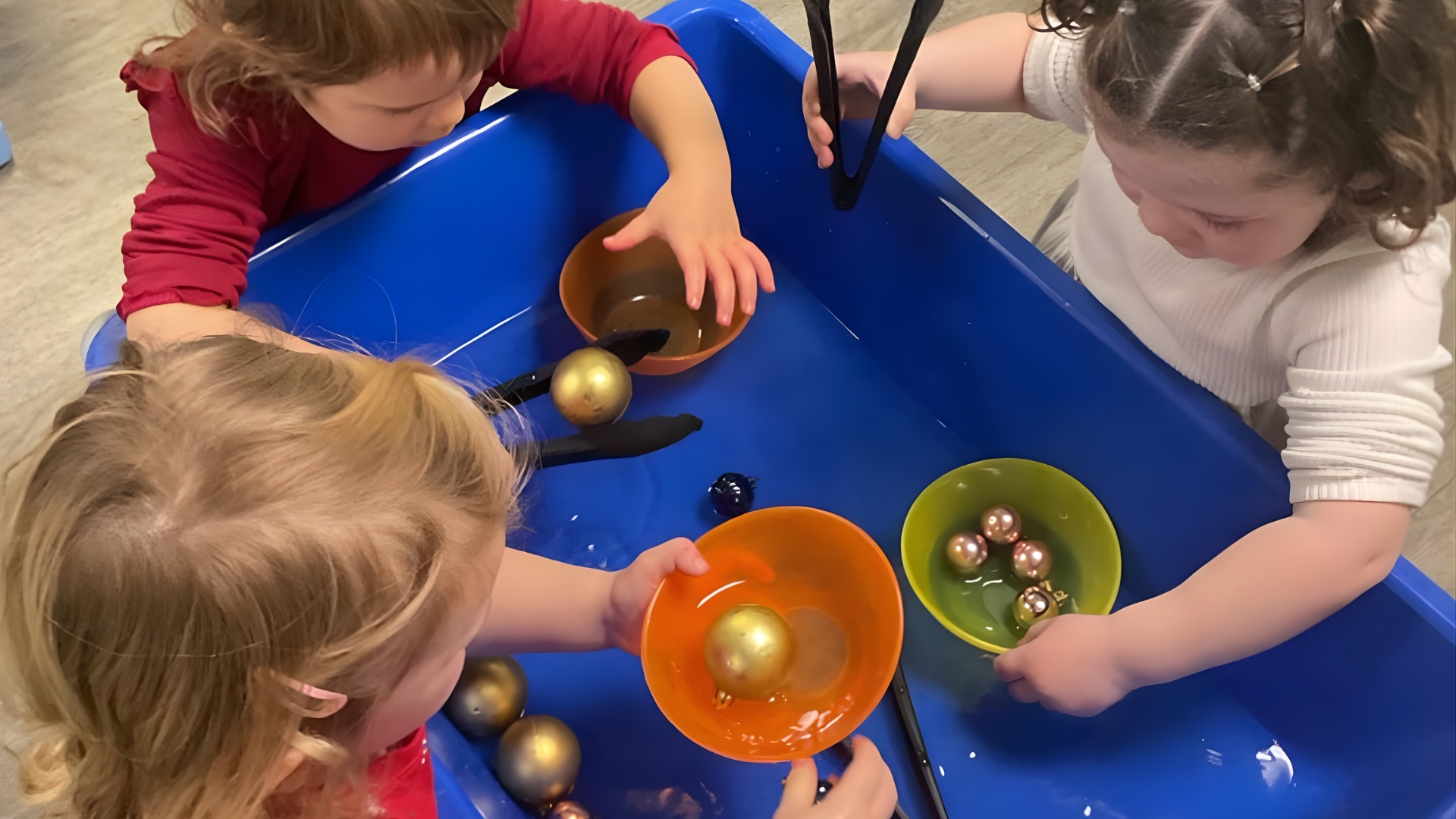 Three children playing with golden balls in colorful bowls inside a blue bin.