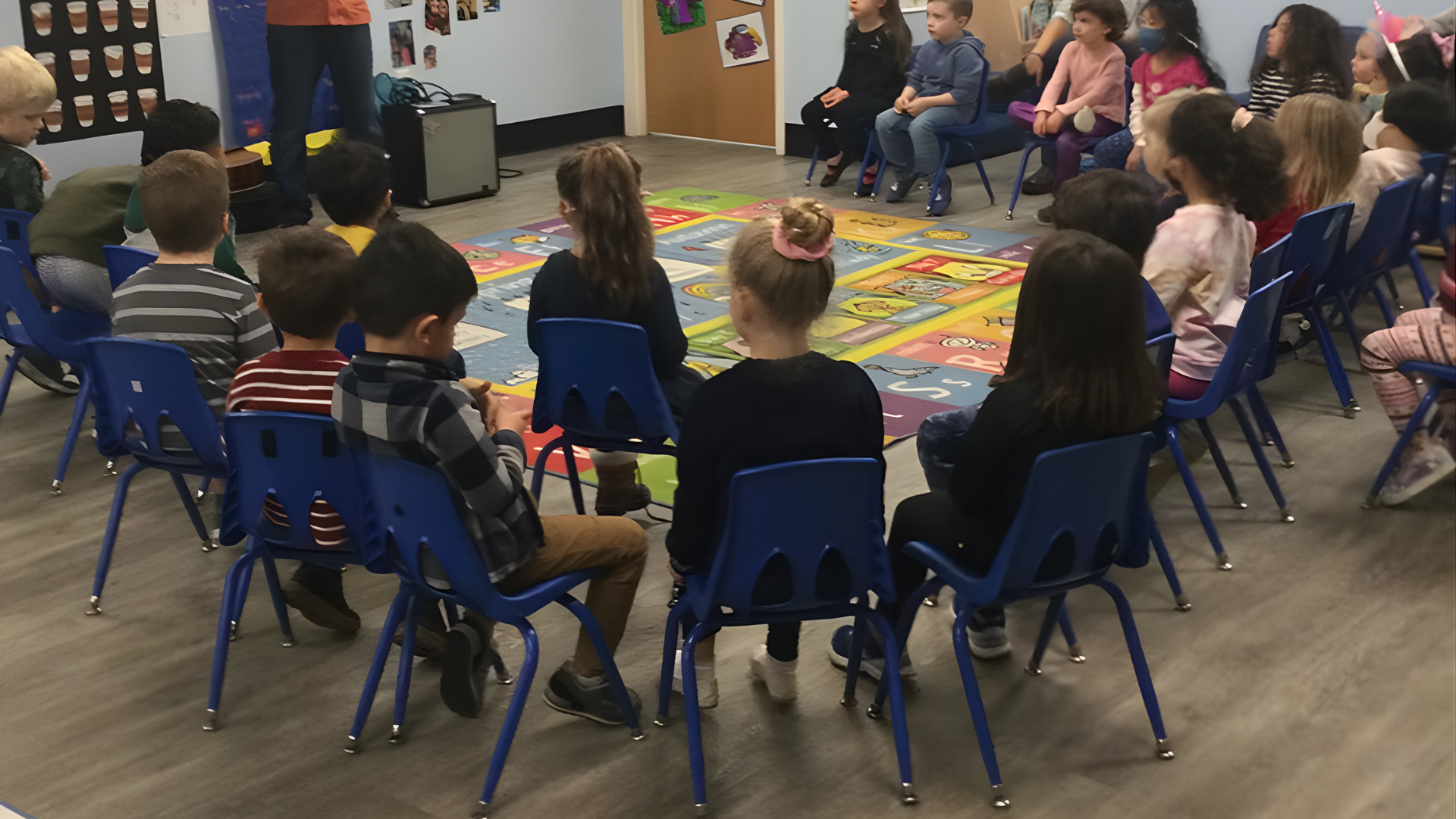 Children sitting in a circle on blue chairs, listening to a person standing in front of them in a classroom.