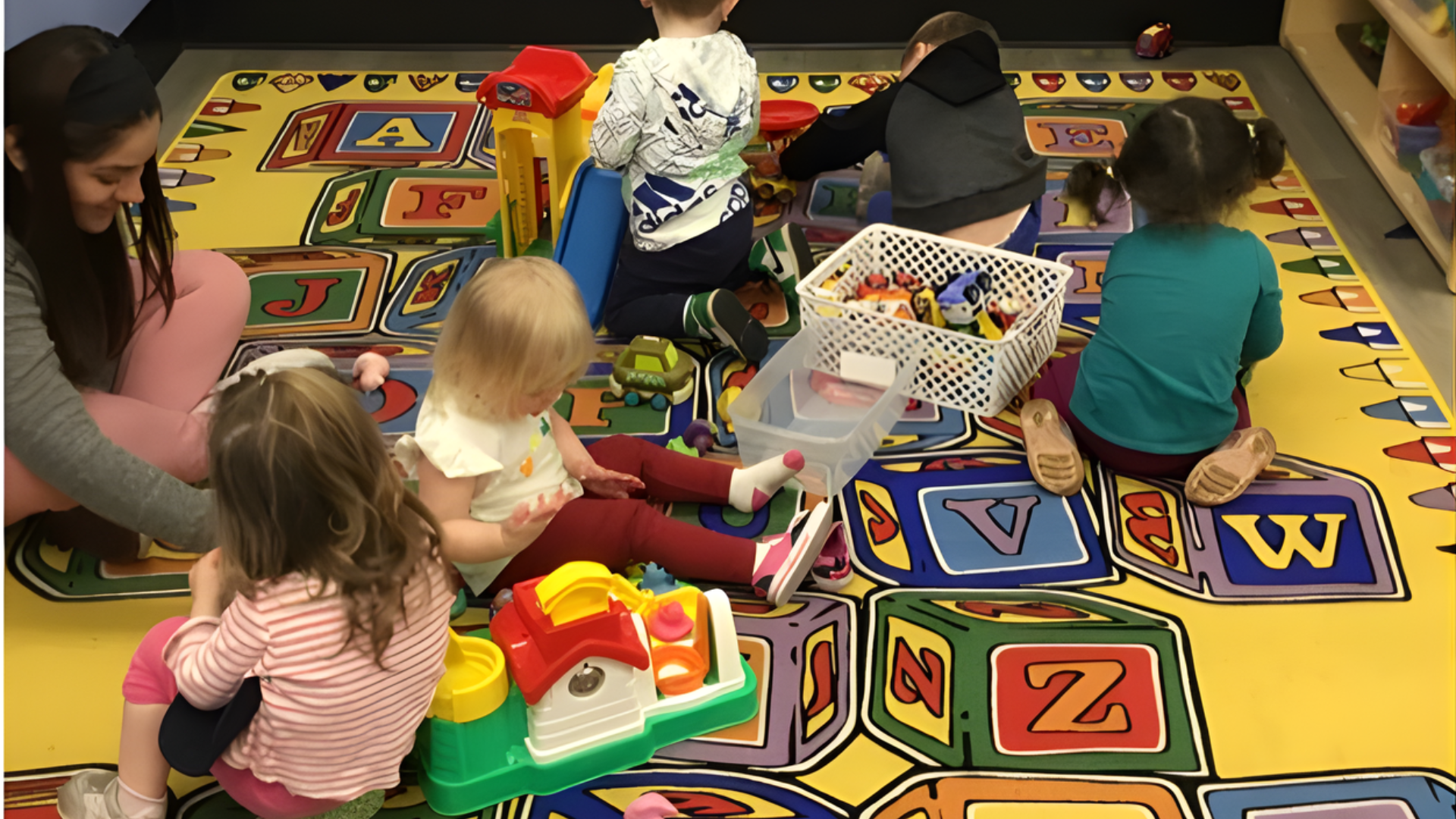 Children and adult playing with toys on a colorful alphabet-themed rug in a playroom.