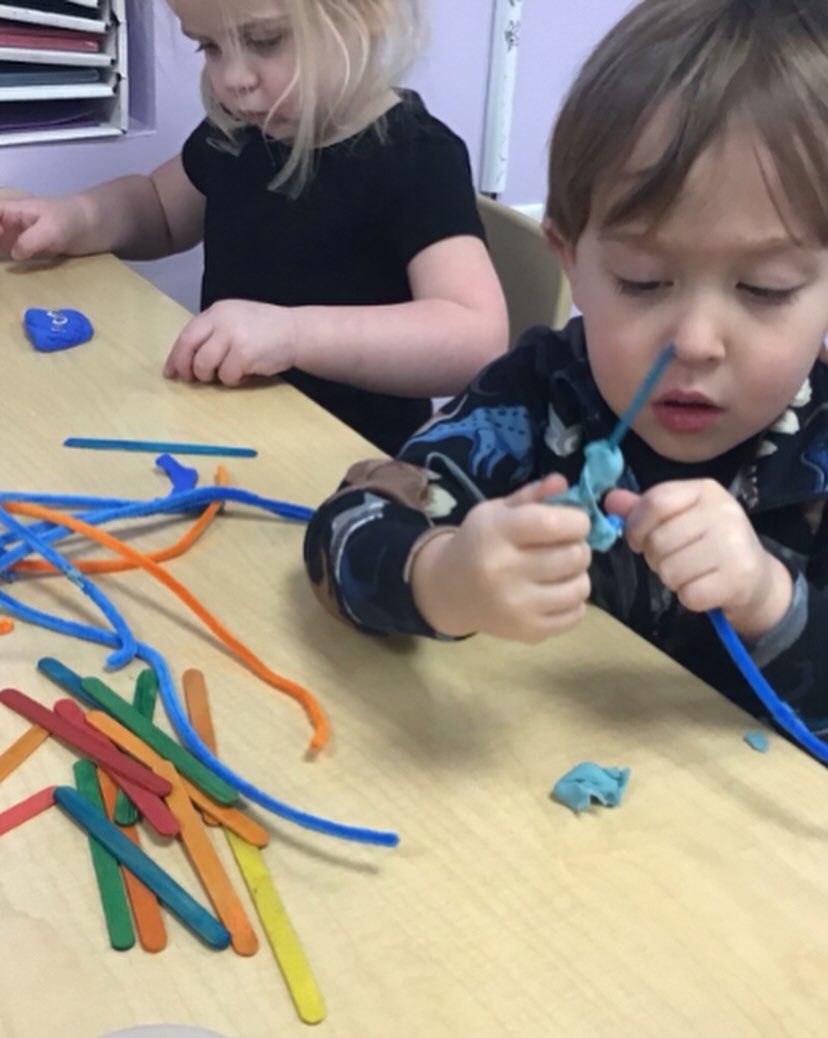 Two young children crafting with playdough, pipe cleaners, and popsicle sticks at a table.