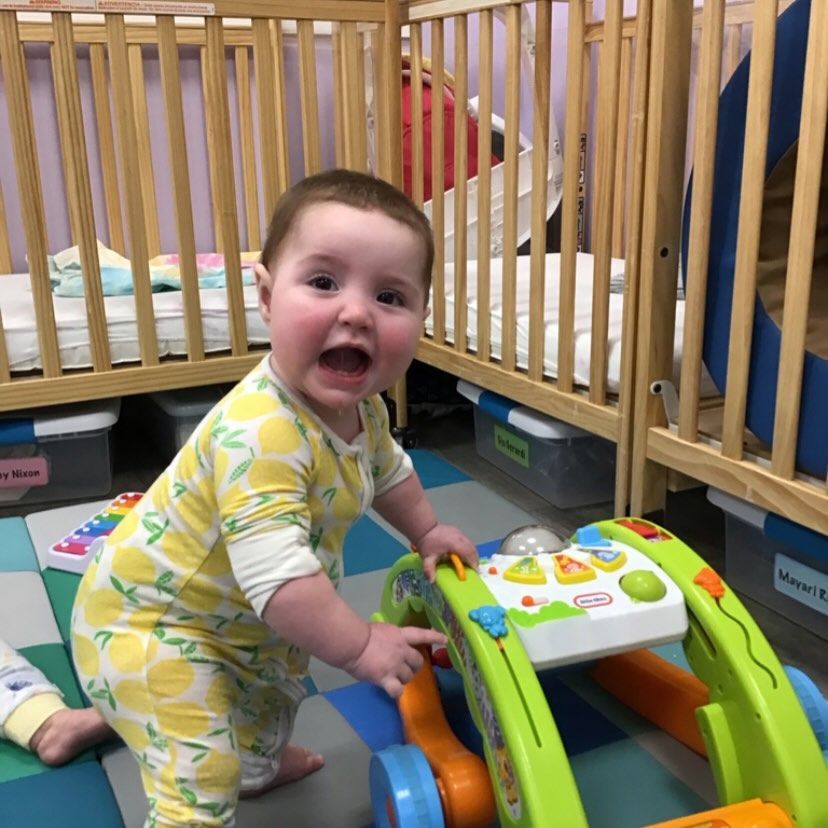 Baby in yellow lemon pajamas plays with a walker in a nursery.