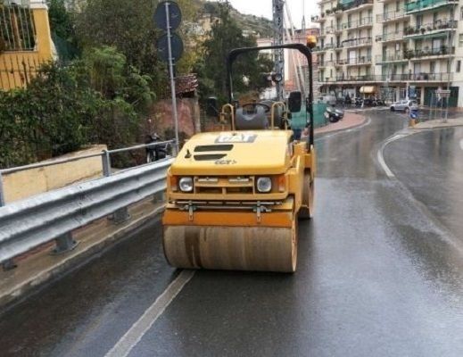 Rullo compressore giallo che compatta l'asfalto su una strada bagnata nei pressi di una zona residenziale.
