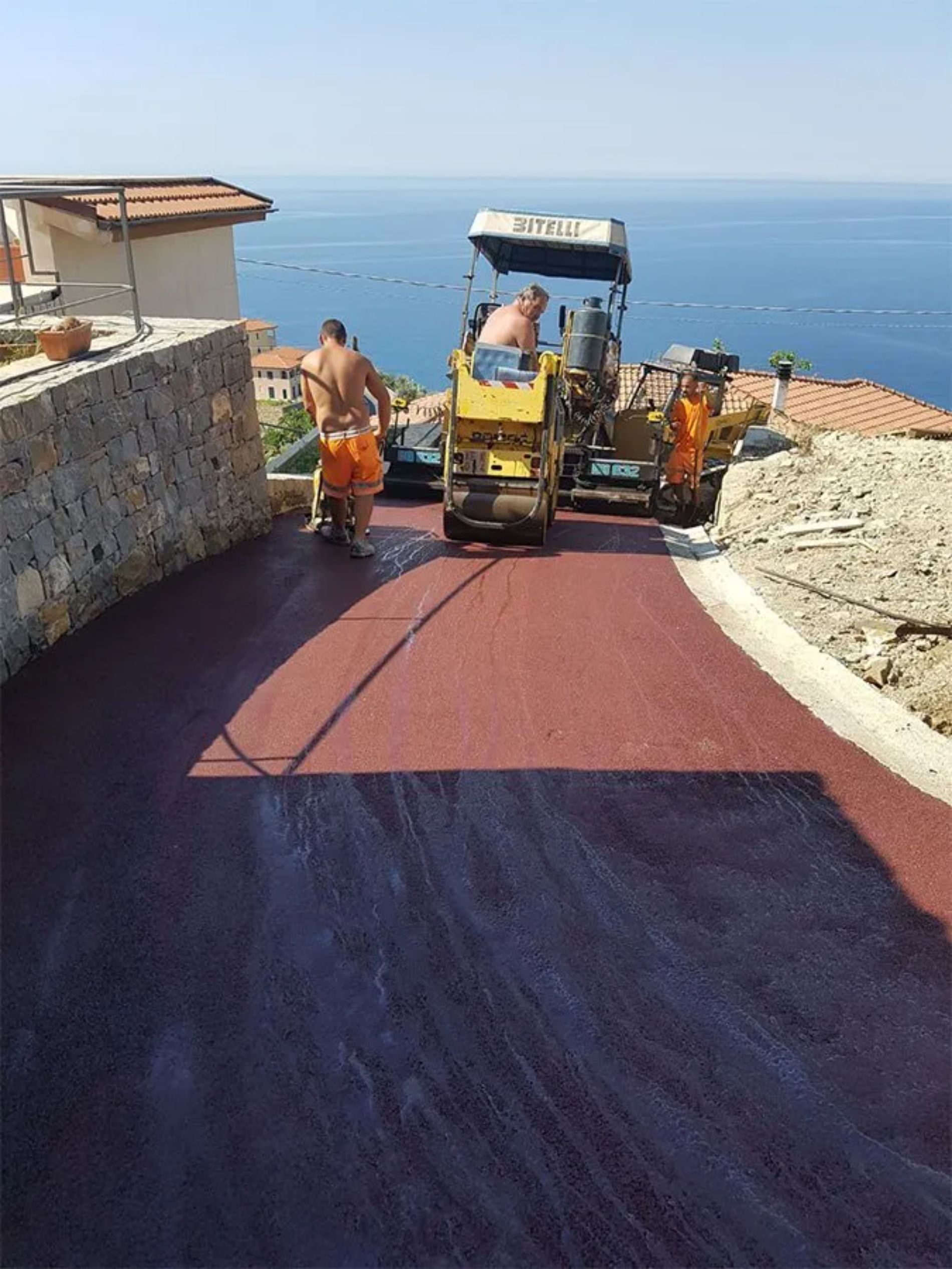 Operai stradali asfaltano una strada rosso-marrone vicino all'oceano. Uno aziona una macchina; un altro usa una piastra vibrante.