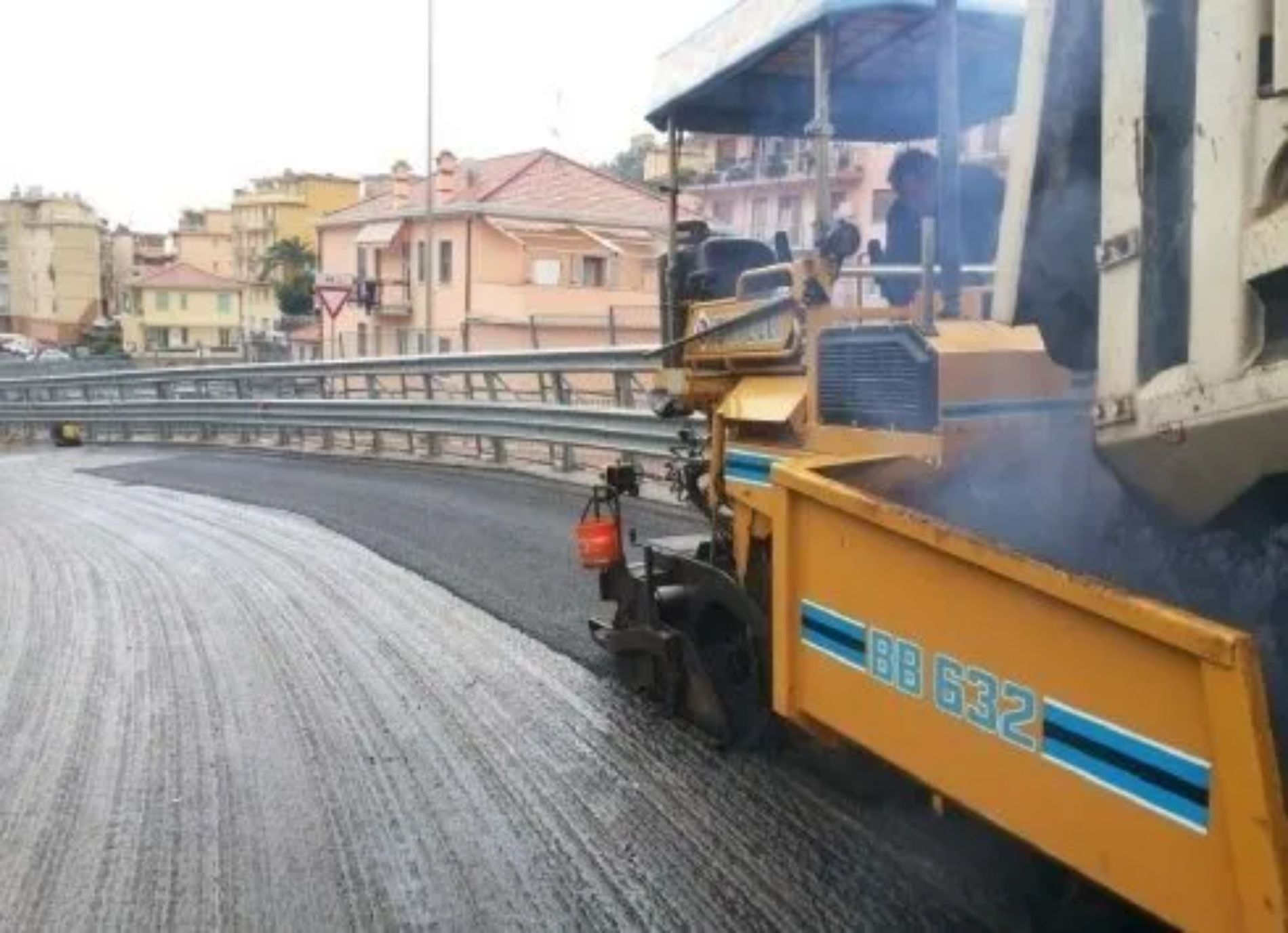 Macchina asfaltatrice che stende uno strato fresco su una strada curva, con edifici sullo sfondo.
