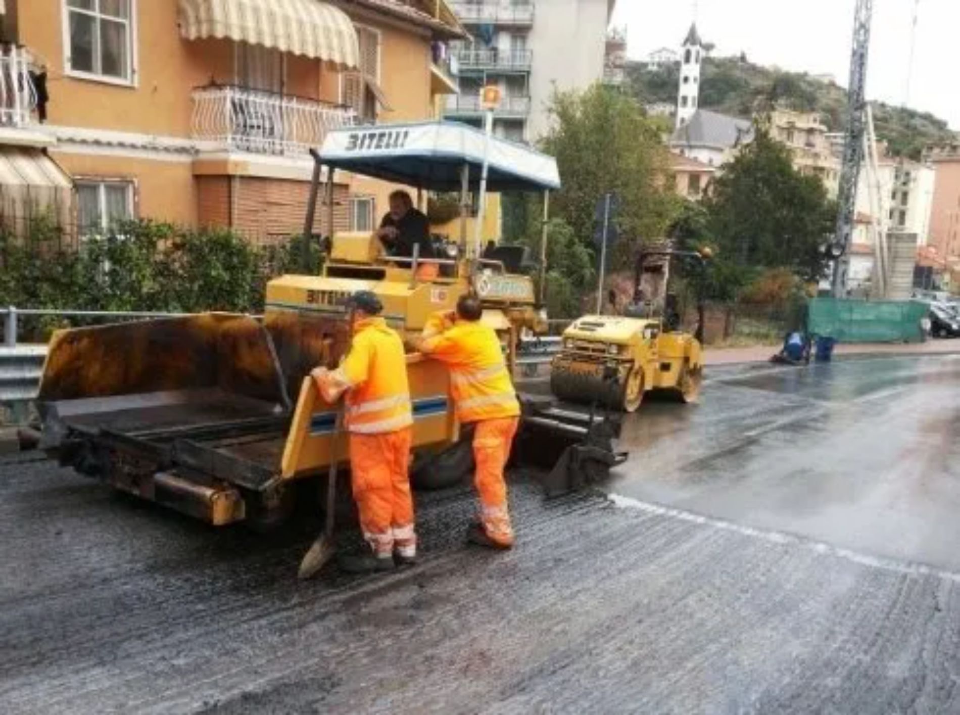 Costruzione di strade: operai con gilet arancioni, macchina asfaltatrice, rullo e contesto urbano.