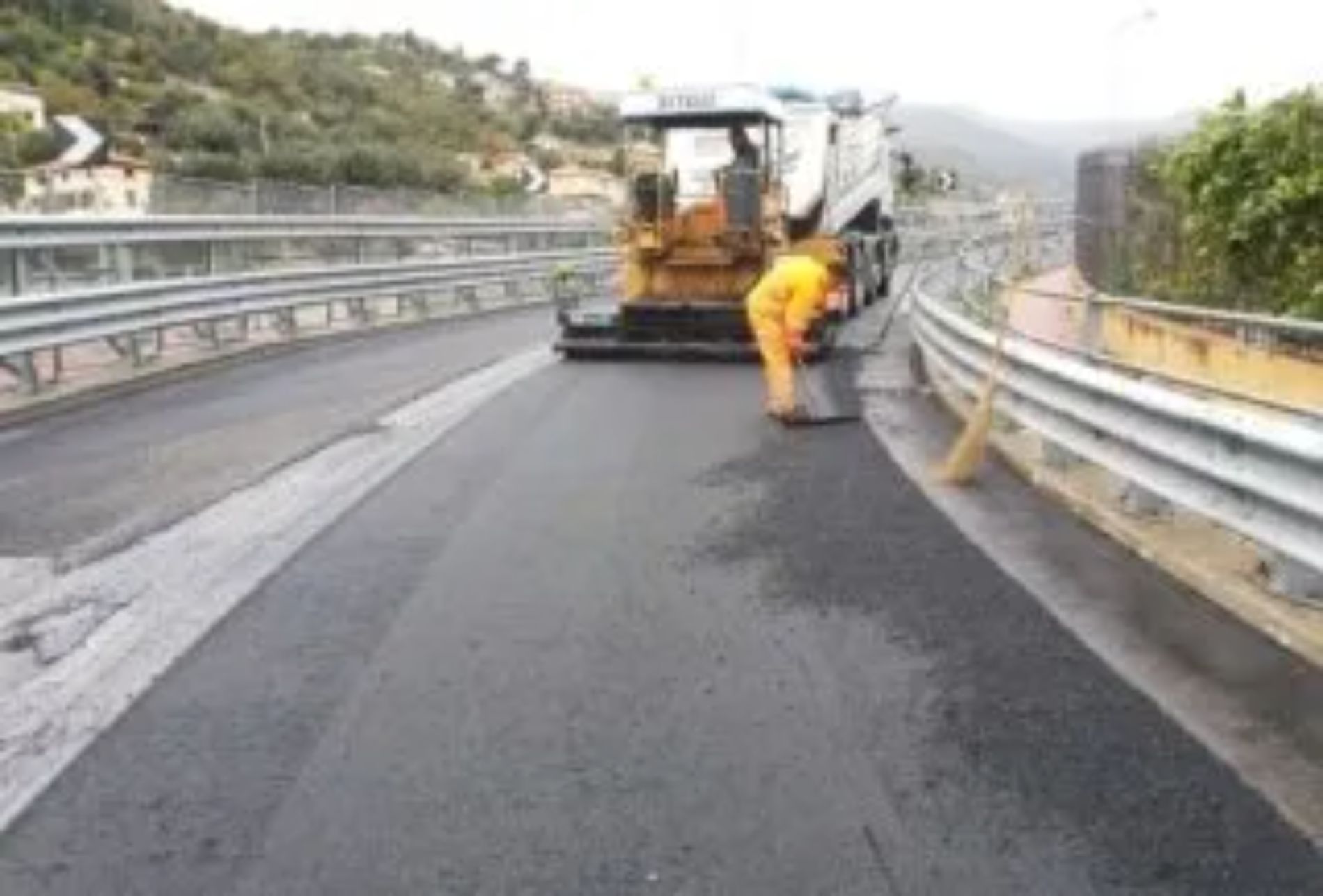 Rifacimento del manto stradale su un ponte; operaio in tuta arancione guida la macchina finitrice.