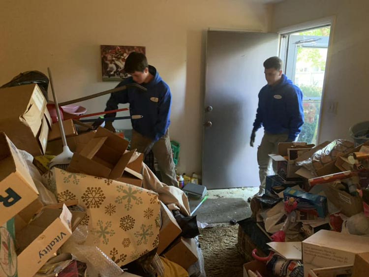 Two men are standing in a room filled with boxes and a box that says weekly