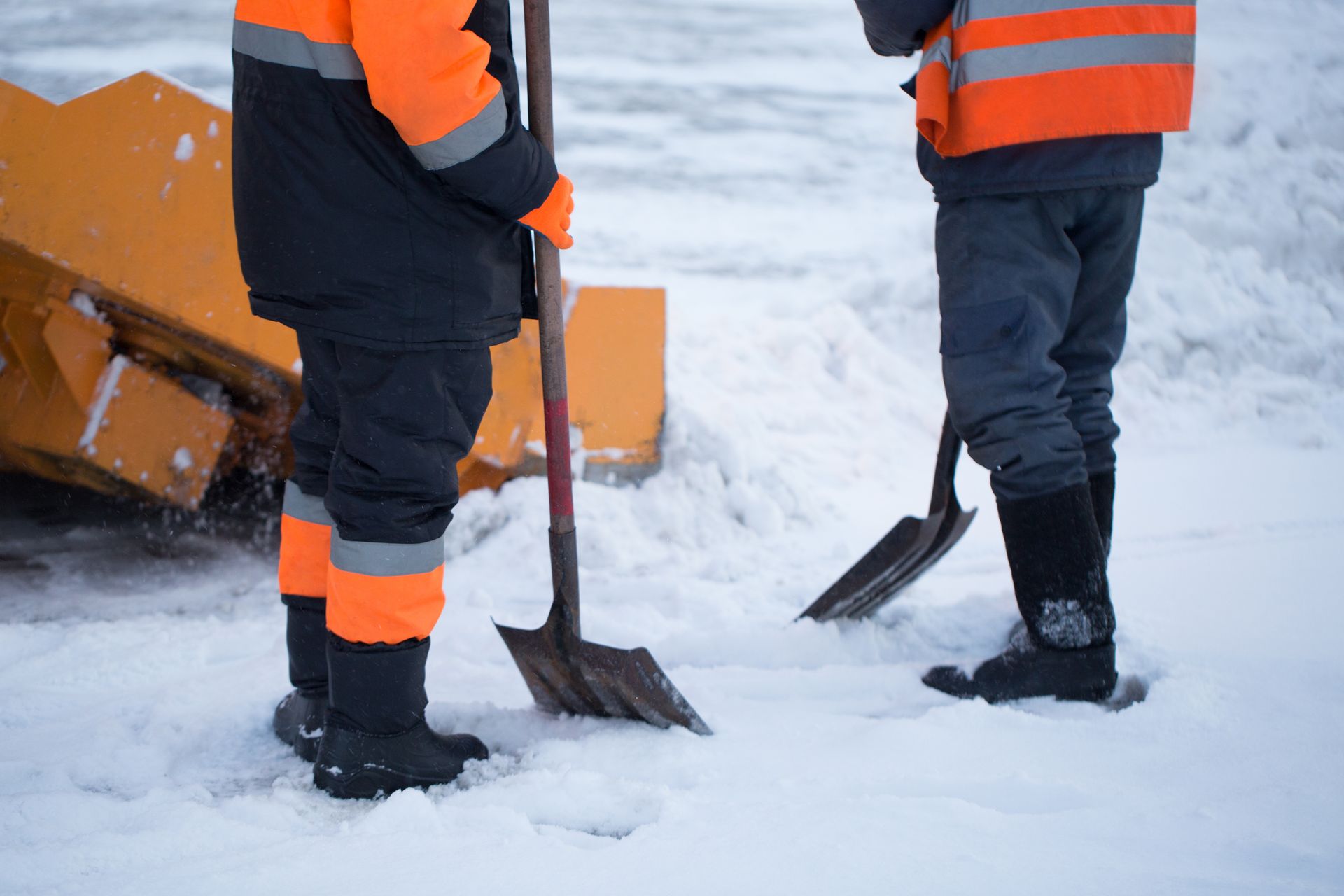 Two workers in orange vests and dark winter gear shovel snow.