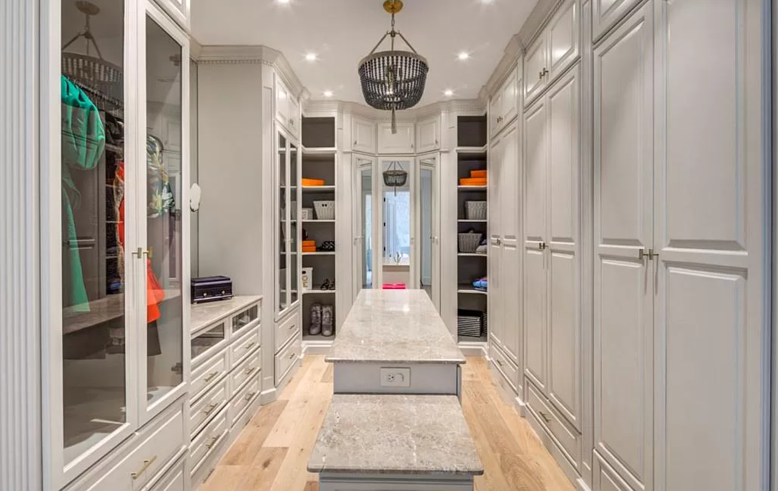 Walk-in closet with pale gray cabinets, marble-topped island, and crystal chandelier.