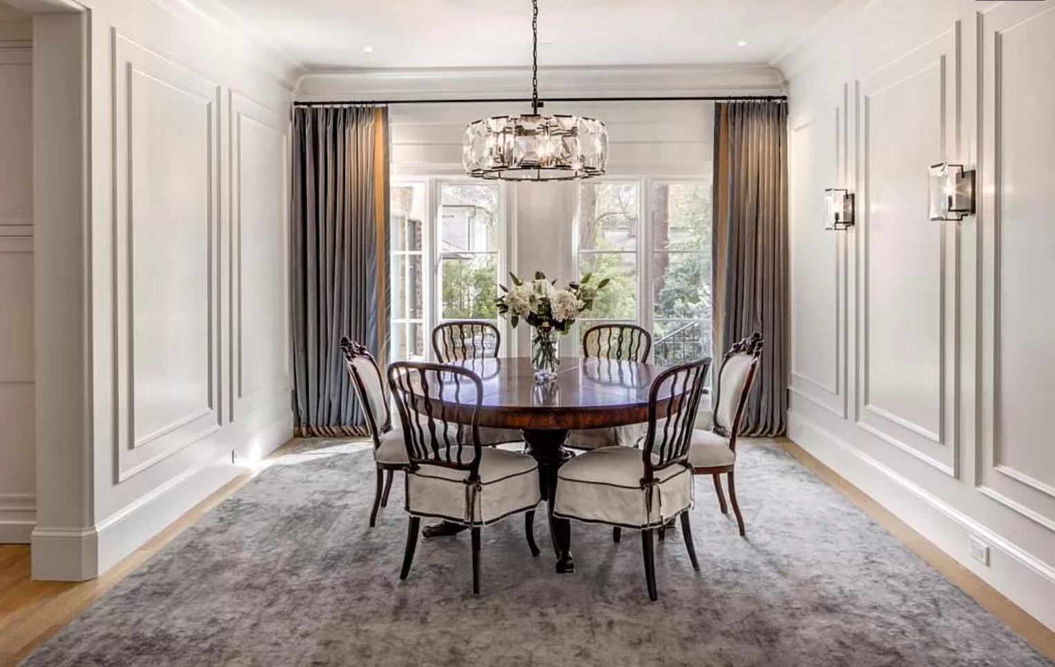 Elegant dining room with round table, chairs, and large window with gray curtains; rug and chandelier.