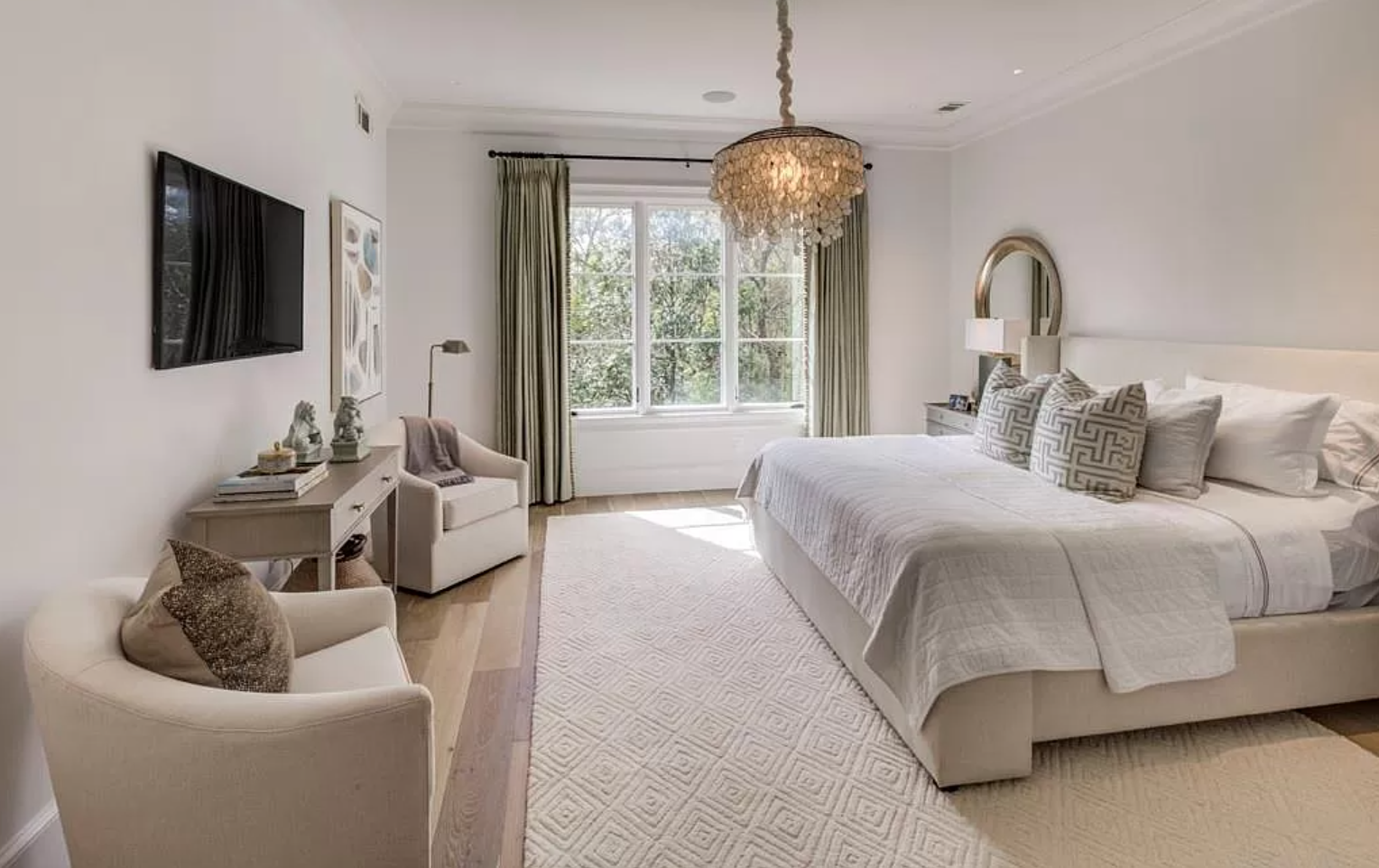 Bedroom with large bed, chandelier, armchairs, and a TV on the wall.