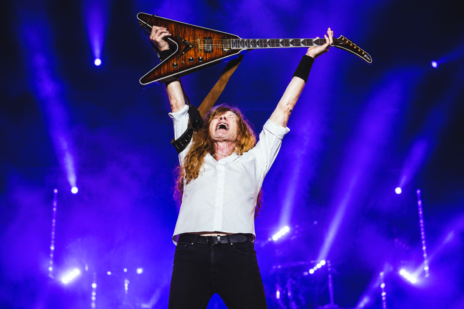 Dave Mustaine raises his arms on stage beneath warm orange-red lights during a live Megadeth performance, with the band's iconic backdrop behind him.
