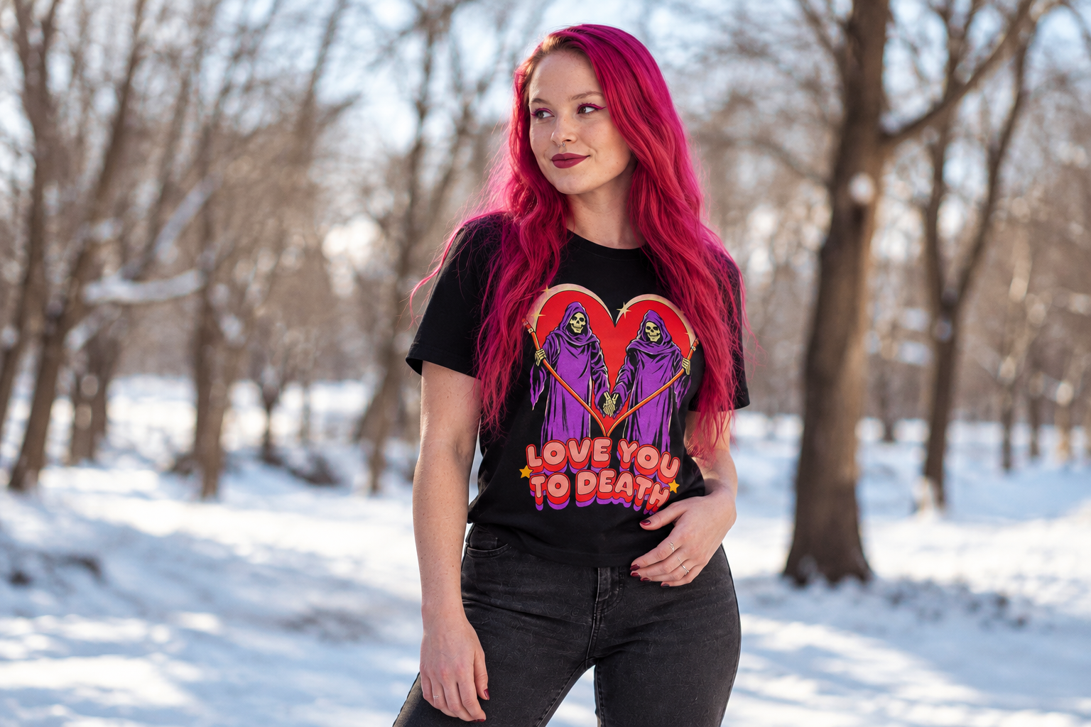 Woman with bright magenta hair wearing a black Love You to Death graphic T-shirt featuring two colourful Grim Reapers in a snowy woodland.