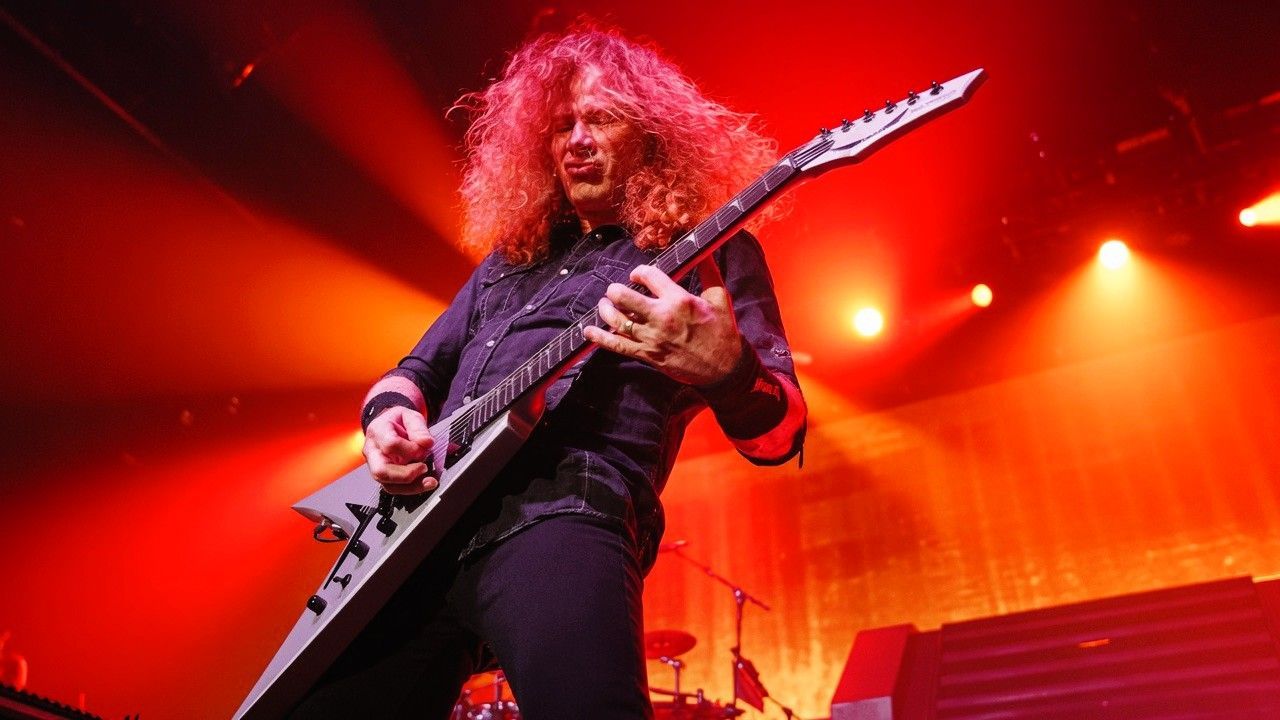 Dave Mustaine performs live under fiery red and orange stage lights during Megadeth's farewell tour, playing his signature Flying V guitar.