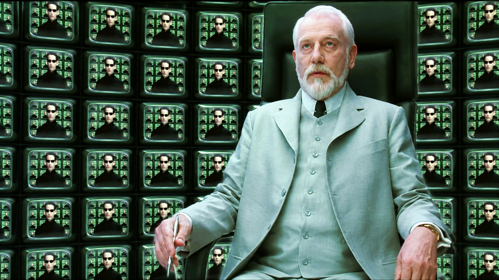 The Architect seated before a wall of screens displaying Neo in The Matrix Reloaded.