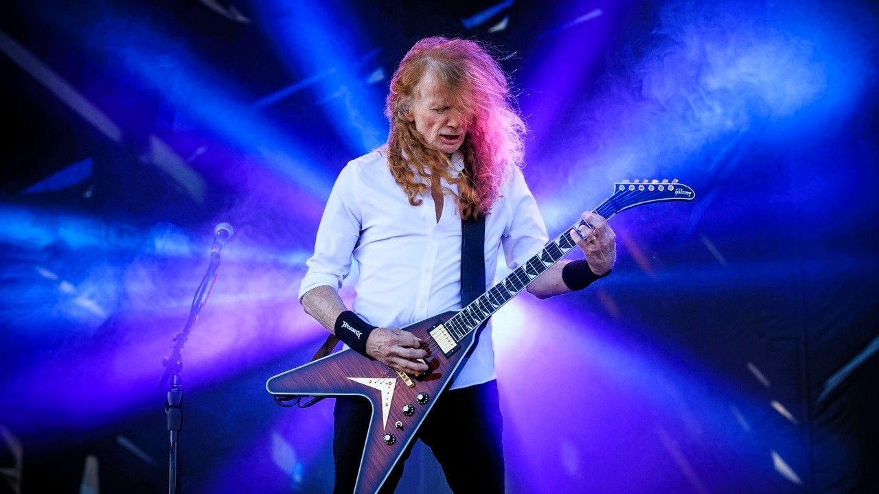 Dave Mustaine performing live on stage with his Flying V guitar during Megadeth's farewell tour beneath dramatic blue and purple concert lighting.