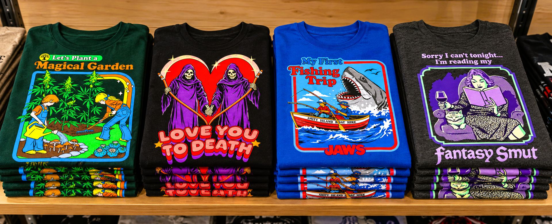Four stacks of colourful graphic T-shirts featuring Magical Garden, Love You to Death, Jaws fishing artwork and Fantasy Smut designs.