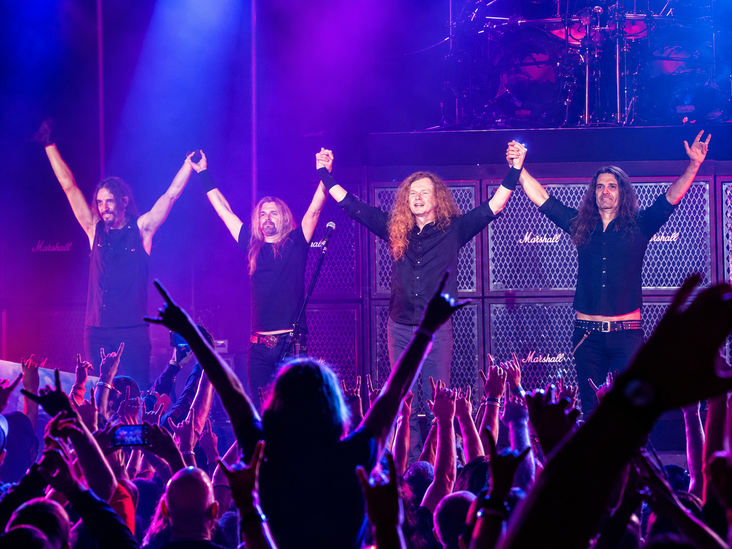 Megadeth take their final bow before a cheering crowd, with Dave Mustaine and the band raising their joined hands beneath vibrant purple, pink and blue stage lights.