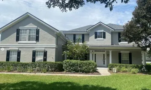 Large gray two-story suburban house with white trim, front porch, and a green lawn under trees