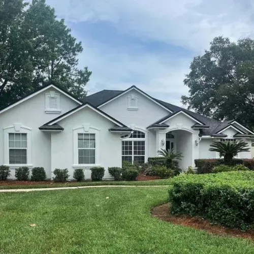 White suburban house with gabled roof, front lawn, shrubs, and trees under a cloudy sky