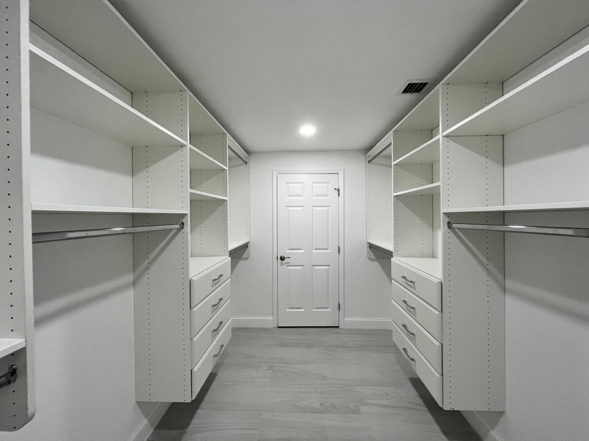 Walk-in closet with white shelving, drawers, and hanging rods on both sides, leading to a closed door.