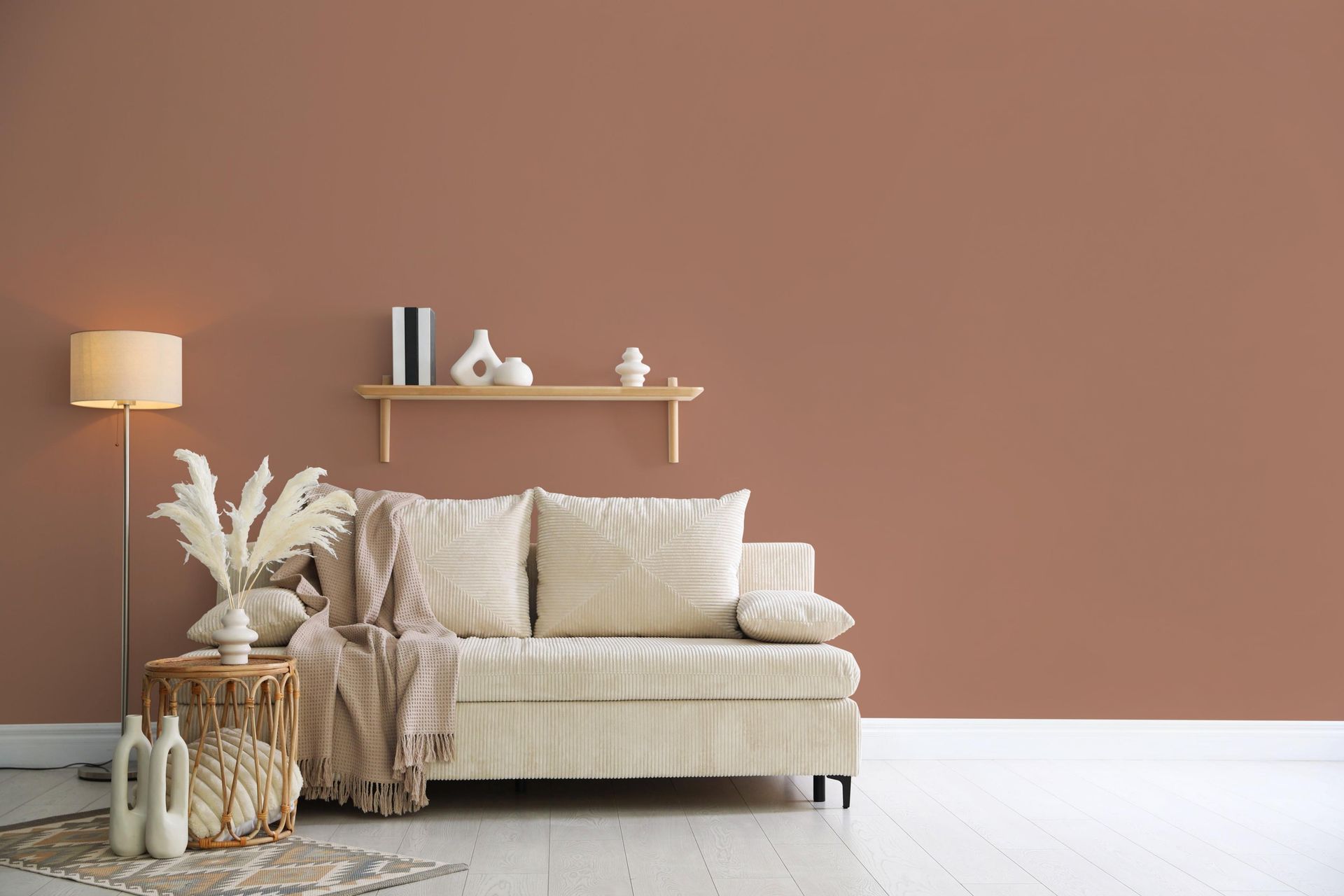 Minimal living room with beige sofa, dried plants, floor lamp, and wood shelf against a terracotta wall