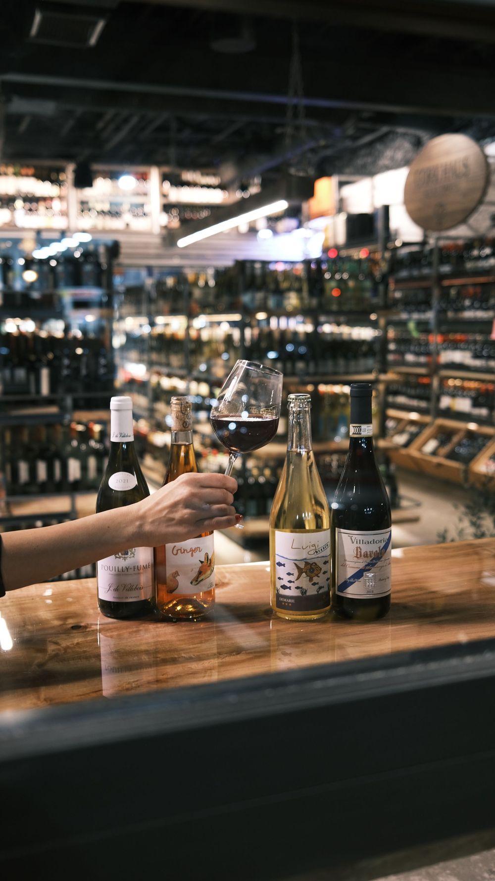 A person is holding a glass of wine while sitting at a table with bottles of wine.