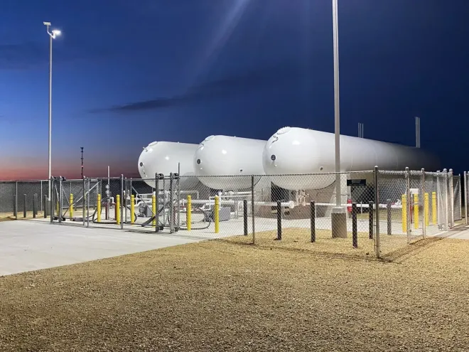 Large white industrial tanks behind a fence at dusk, lit by tall streetlights in a gravel lot