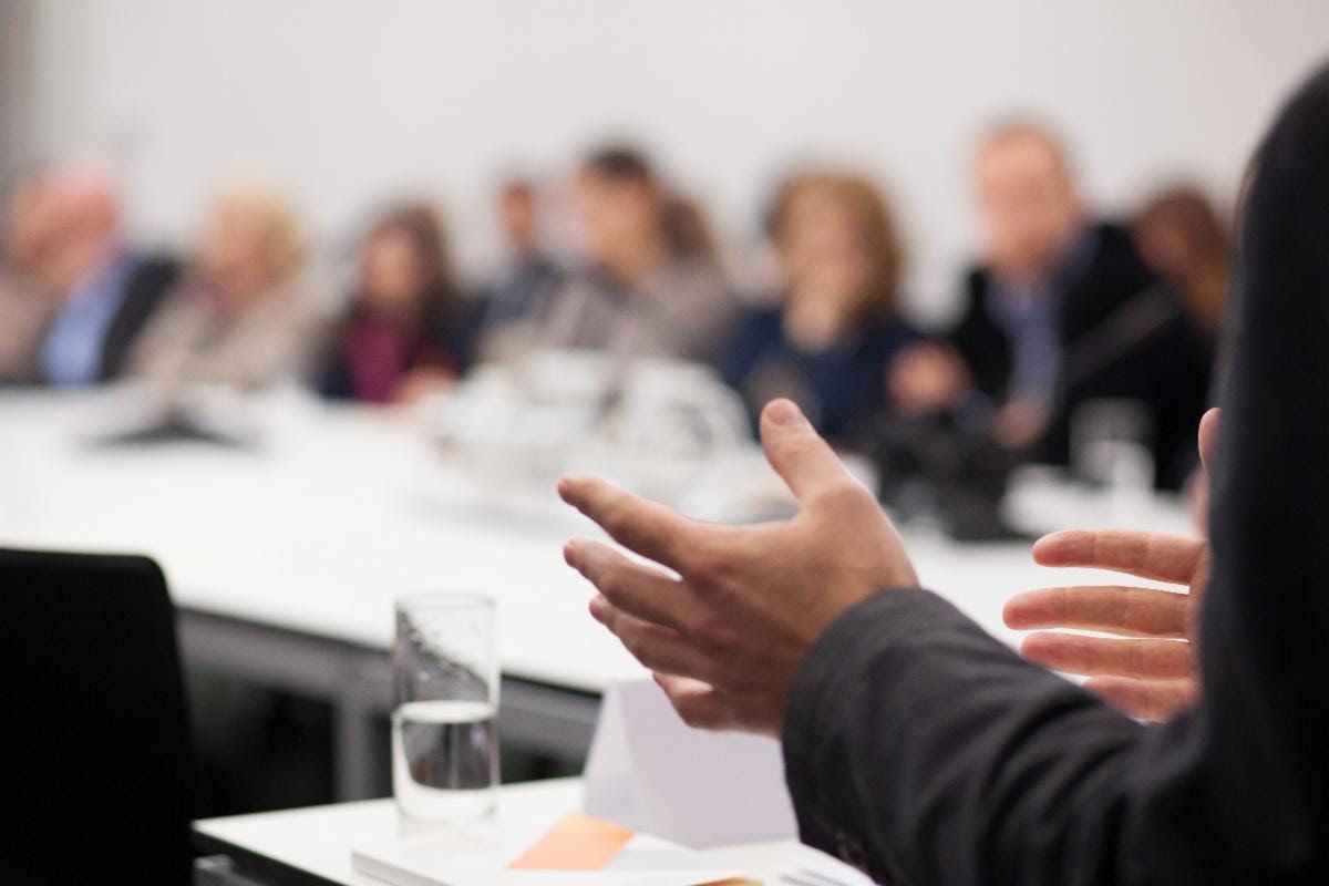 A presenter gestures while speaking to a seated group during a confidential buyer meeting for a busi