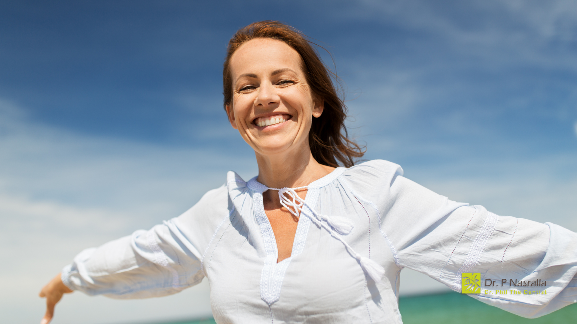 Woman with arms outstretched smiling brightly in front of a blue sky and ocean. She wears a white top.