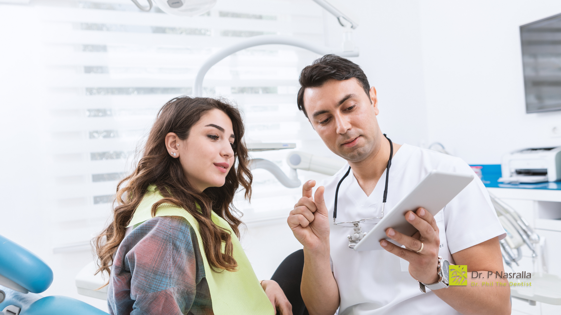 A dentist is talking to a patient in a dental chair while holding a tablet.