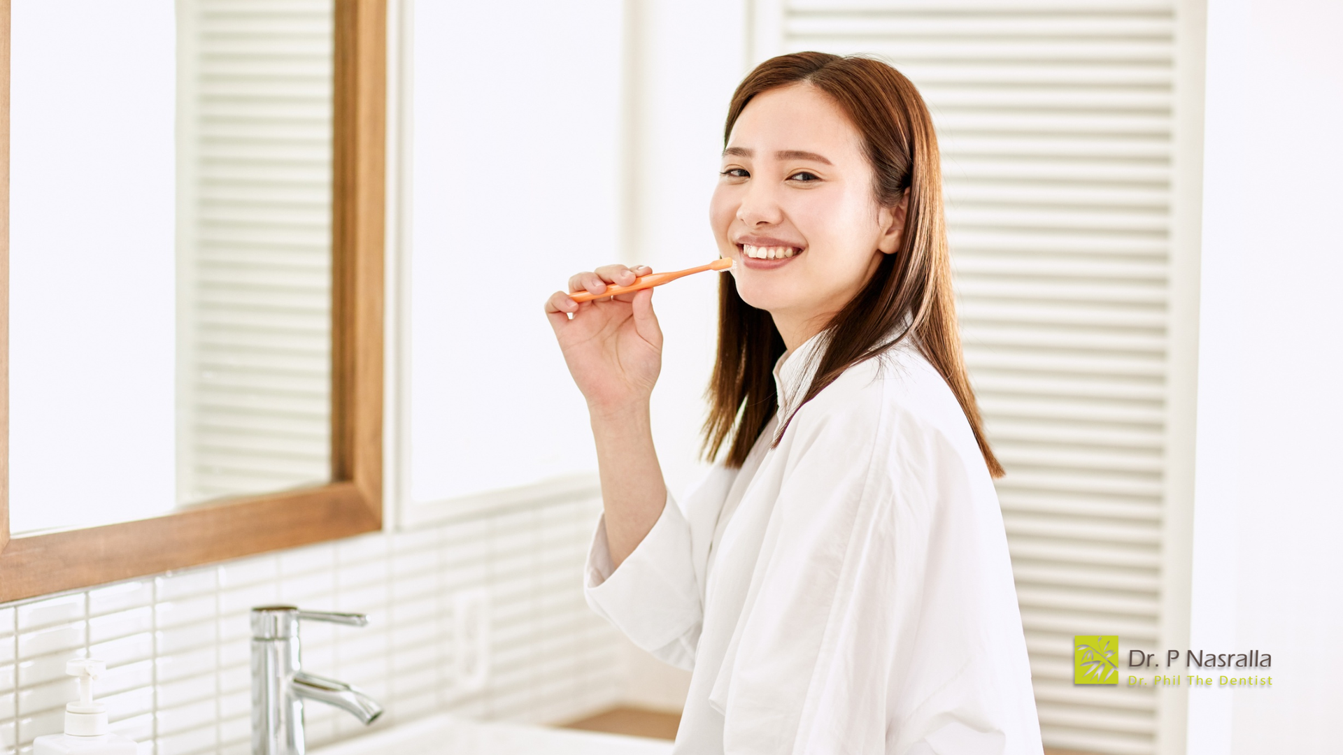 Woman in white robe brushing teeth in a bathroom with a mirror and sink.