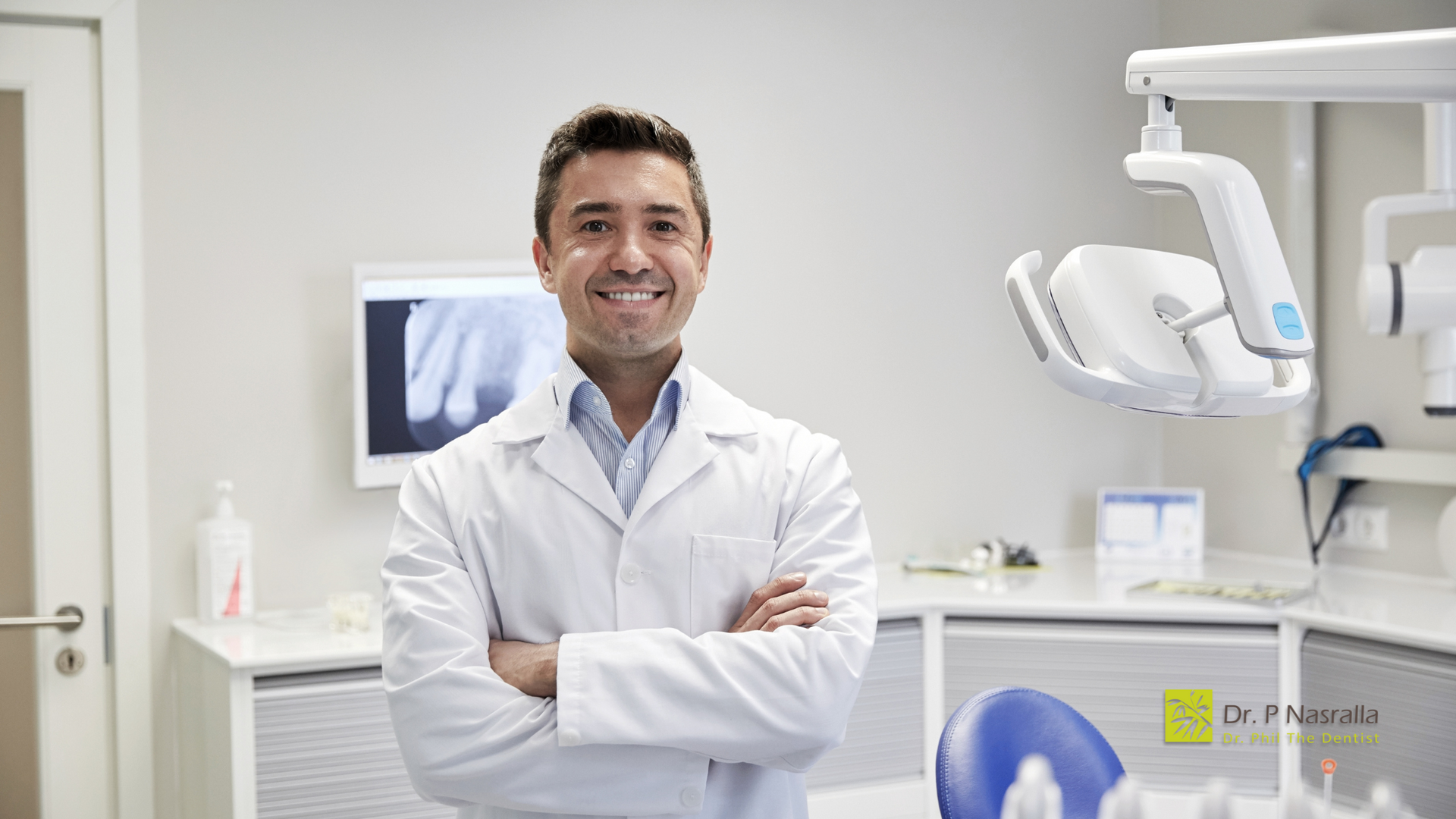 A dentist is standing in a dental office with his arms crossed and smiling.